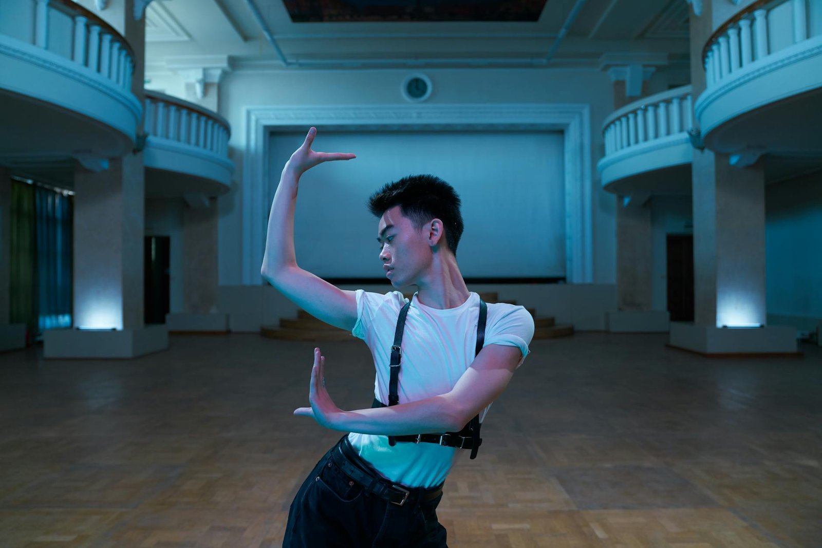 Asian man performs a contemporary dance in a spacious, elegant hall, white shirt and belt straps visible.