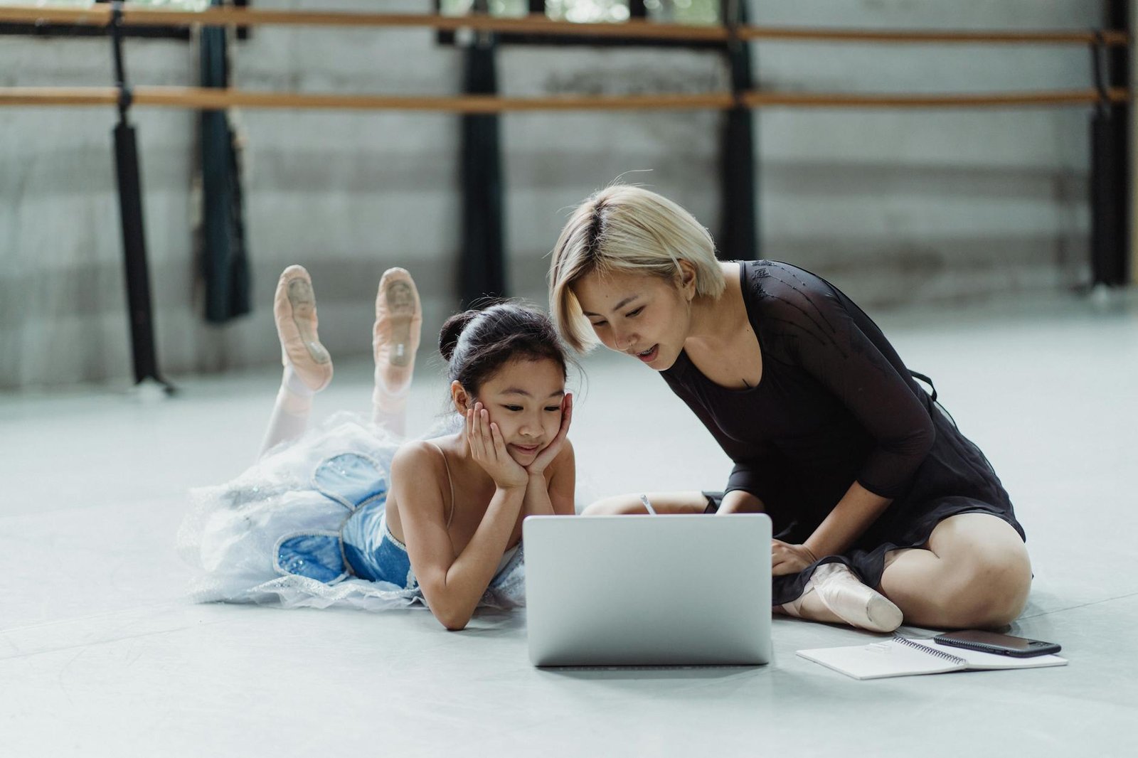 Full body of ethnic little girl wearing tutu lying on floor near personal trainer and watching netbook in dance studio