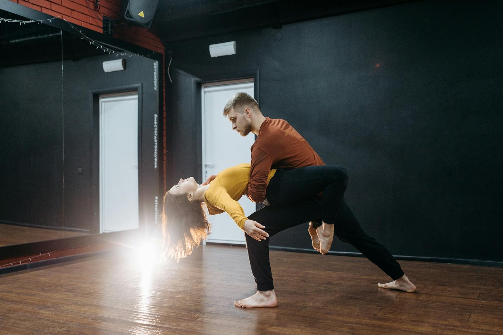 Couple performing an expressive dance move barefoot indoors on a wooden floor.