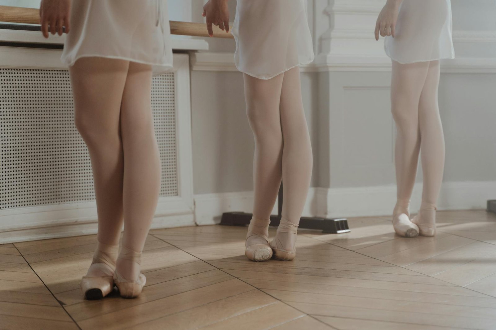 Three young ballerinas in pointe shoes practicing at a barre in a dance studio.