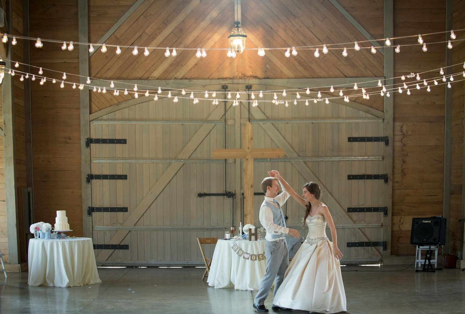 A couple shares a joyful dance amidst twinkling lights in a rustic barn wedding celebration.