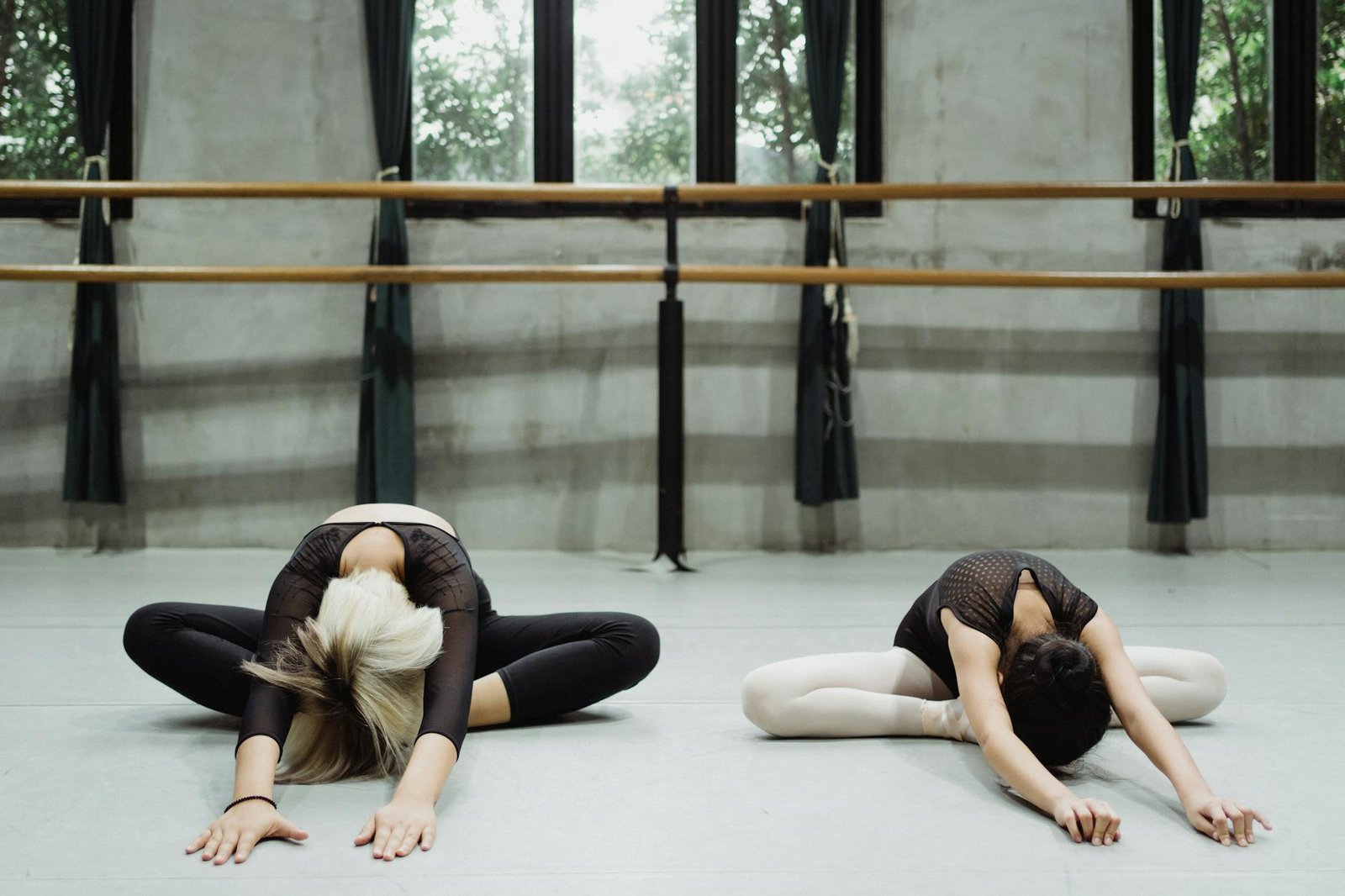 Ballet teacher and ballerina sitting and stretching with bowed heads on floor in studio with barre in daylight