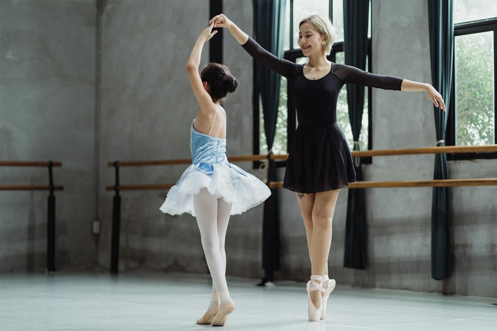 Full body cheerful Asian female ballet instructor standing on tiptoes and dancing with ballerina girl wearing tutu in modern studio