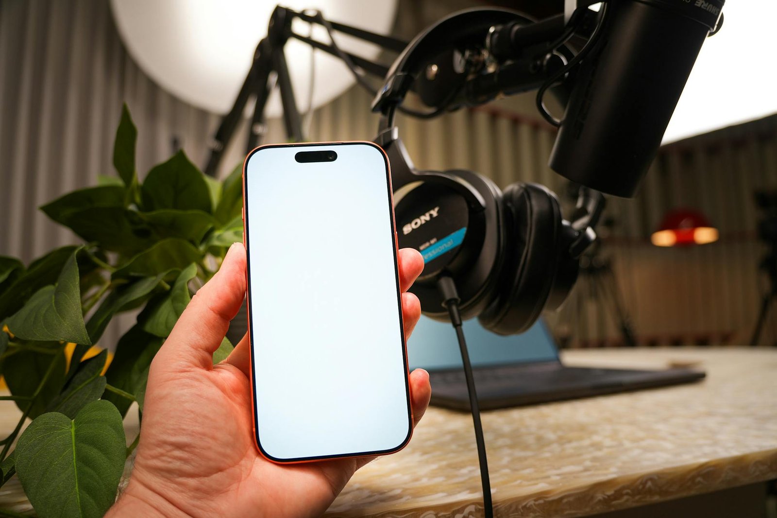 A hand holding a smartphone in a recording studio with microphone and headphones on a desk.