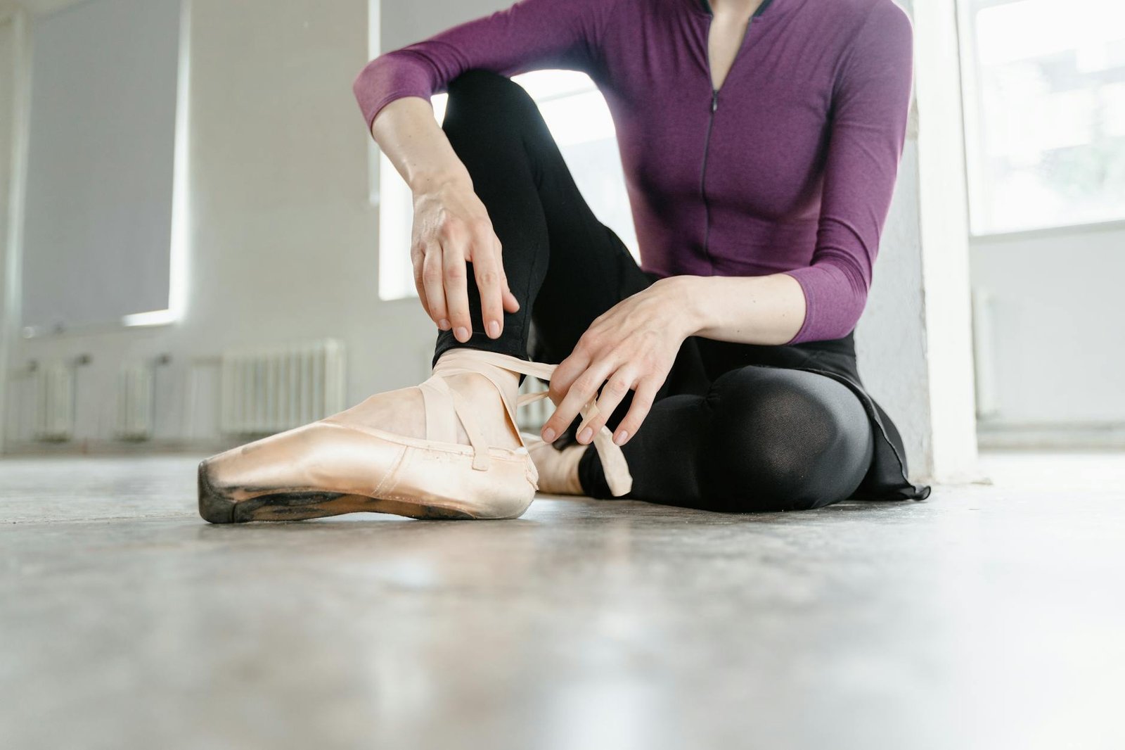 Ballet dancer ties shoes in studio wearing purple top and black leggings.