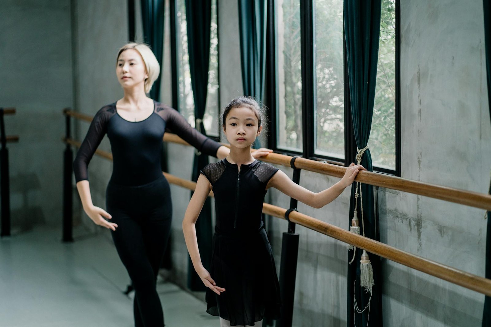 Young ethnic female trainer in uniform teaching pupil near wooden barres in dancing hall while looking away