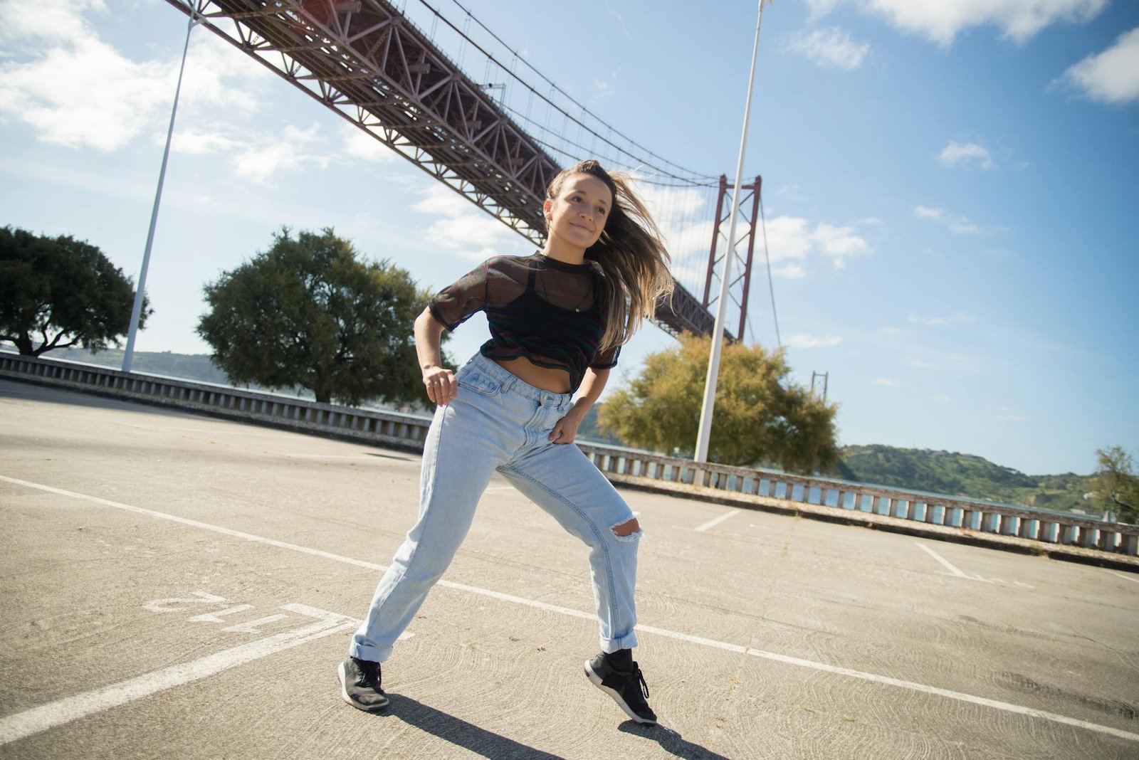 A woman dances energetically beneath the 25 de Abril Bridge in Lisbon, Portugal, showcasing street fashion and urban style.