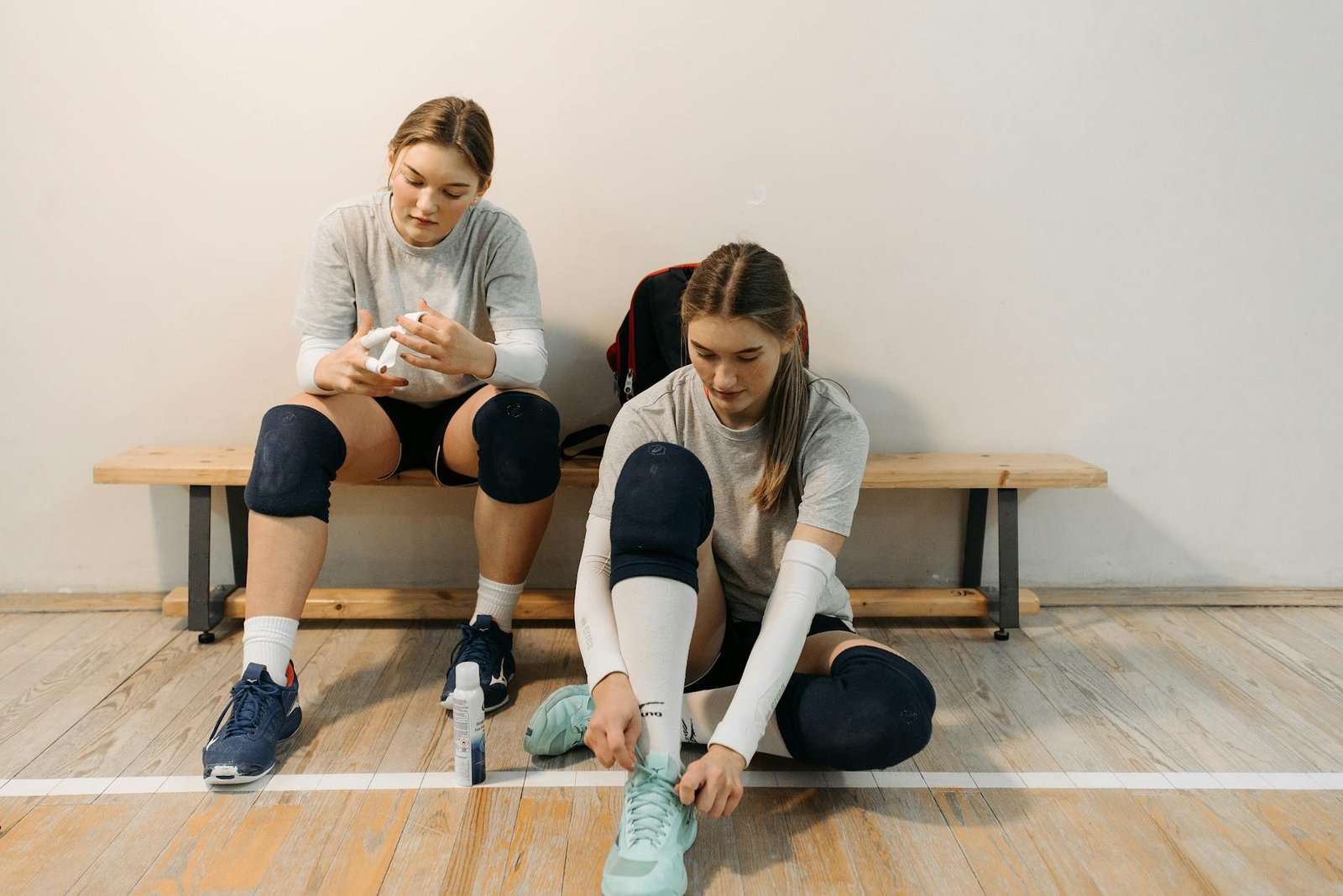 Two female volleyball players in sports gear prepare for a game, sitting on a wooden bench.