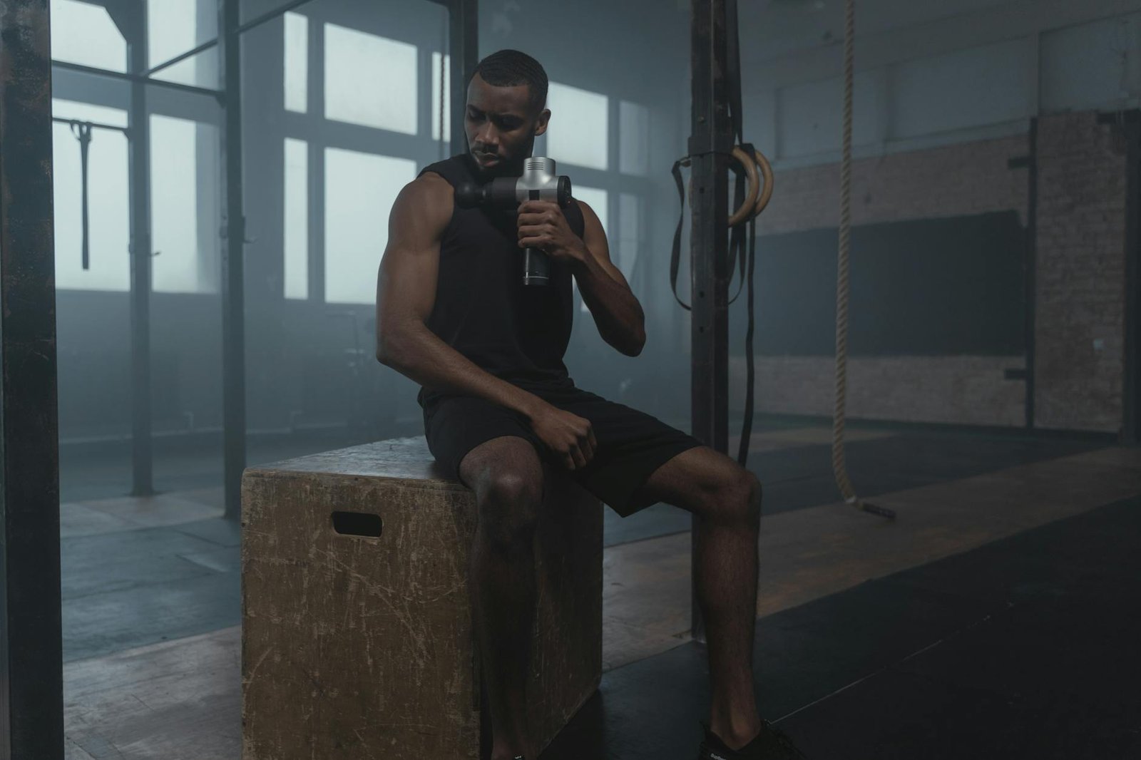 Fit man using a massage gun in a gym, wearing a tank top while seated on a wooden box.