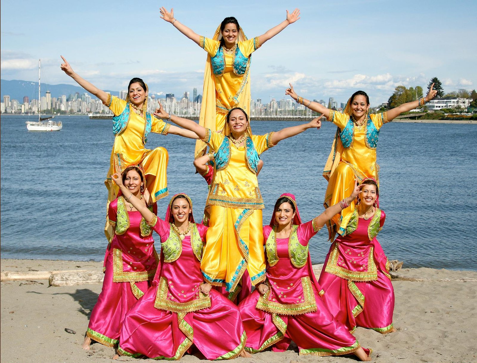 A joyful group of women performs a traditional dance on a sunny beach with a cityscape backdrop.