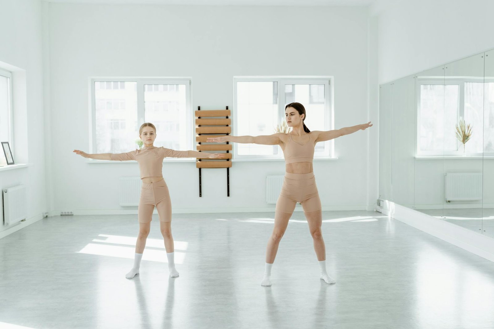 Two women practicing dance poses in a bright, minimalist studio with mirror reflections.