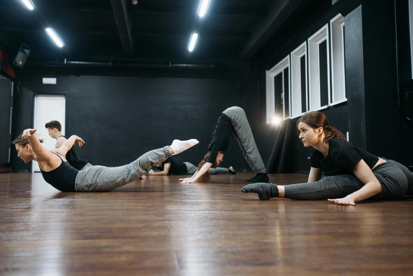 A group of adults practicing dance flexibility exercises together indoors on a wooden floor.