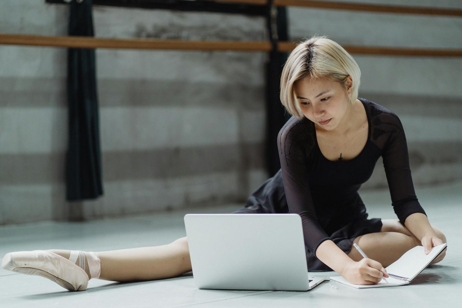Full body of Asian ballerina writing notes in planner while working on laptop in light spacious studio