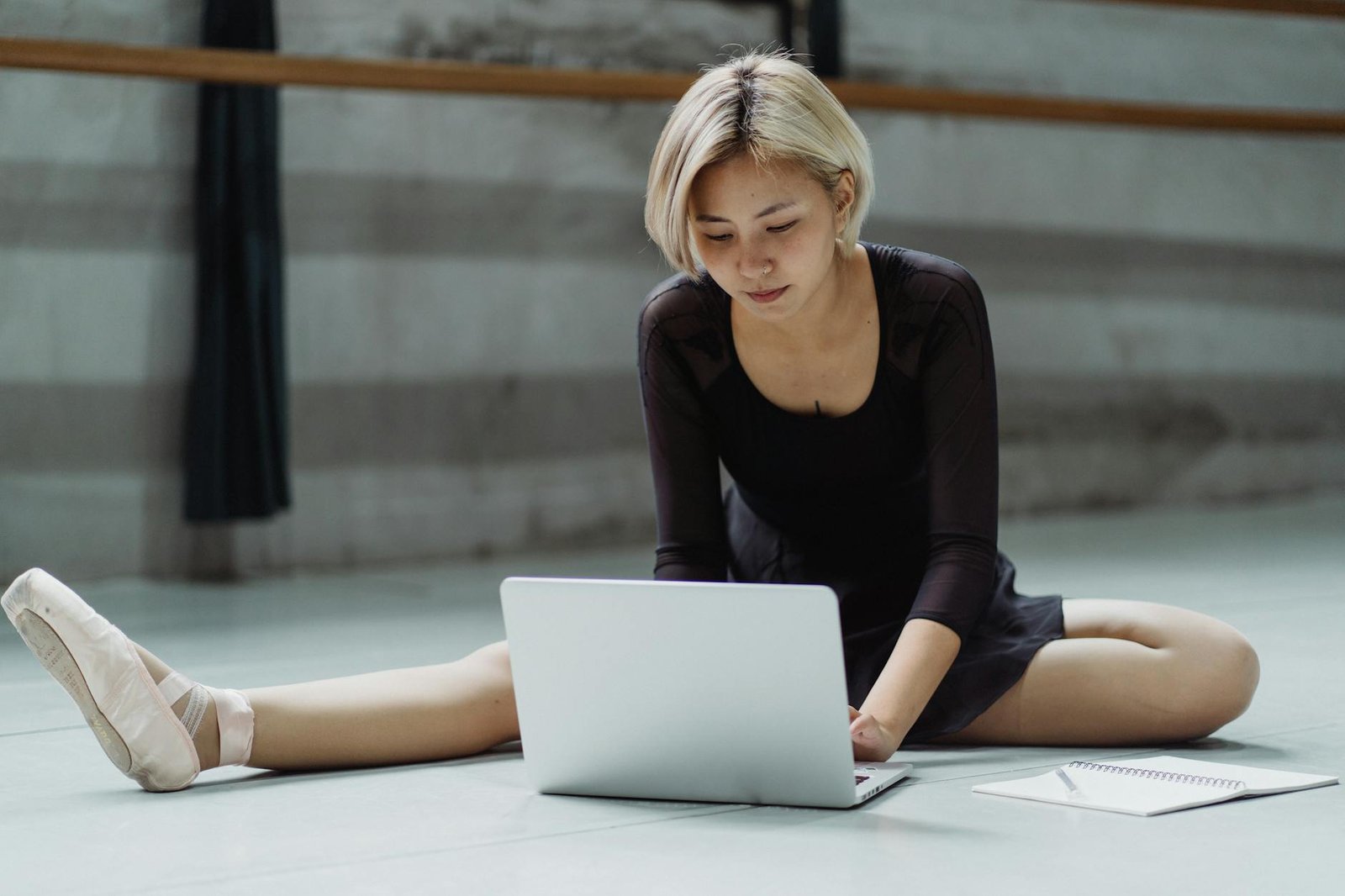 Full body of focused Asian female wearing pointe shoes sitting on floor with tucked leg in studio while typing on laptop