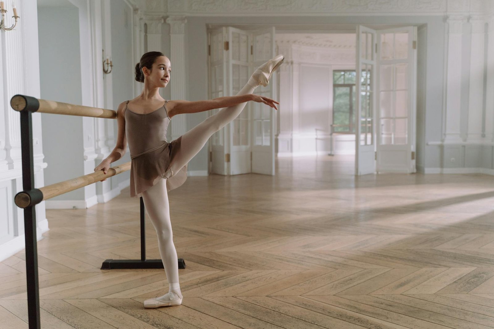A young ballerina gracefully stretches at a barre in a sunlit, elegant dance studio.