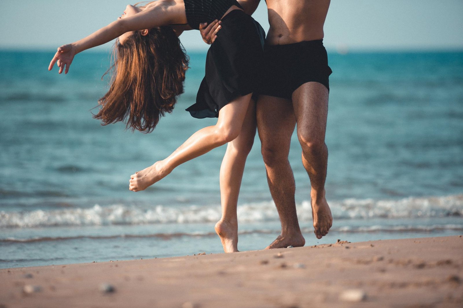 Ballet dancers elegantly perform barefoot on a sandy beach by the ocean.