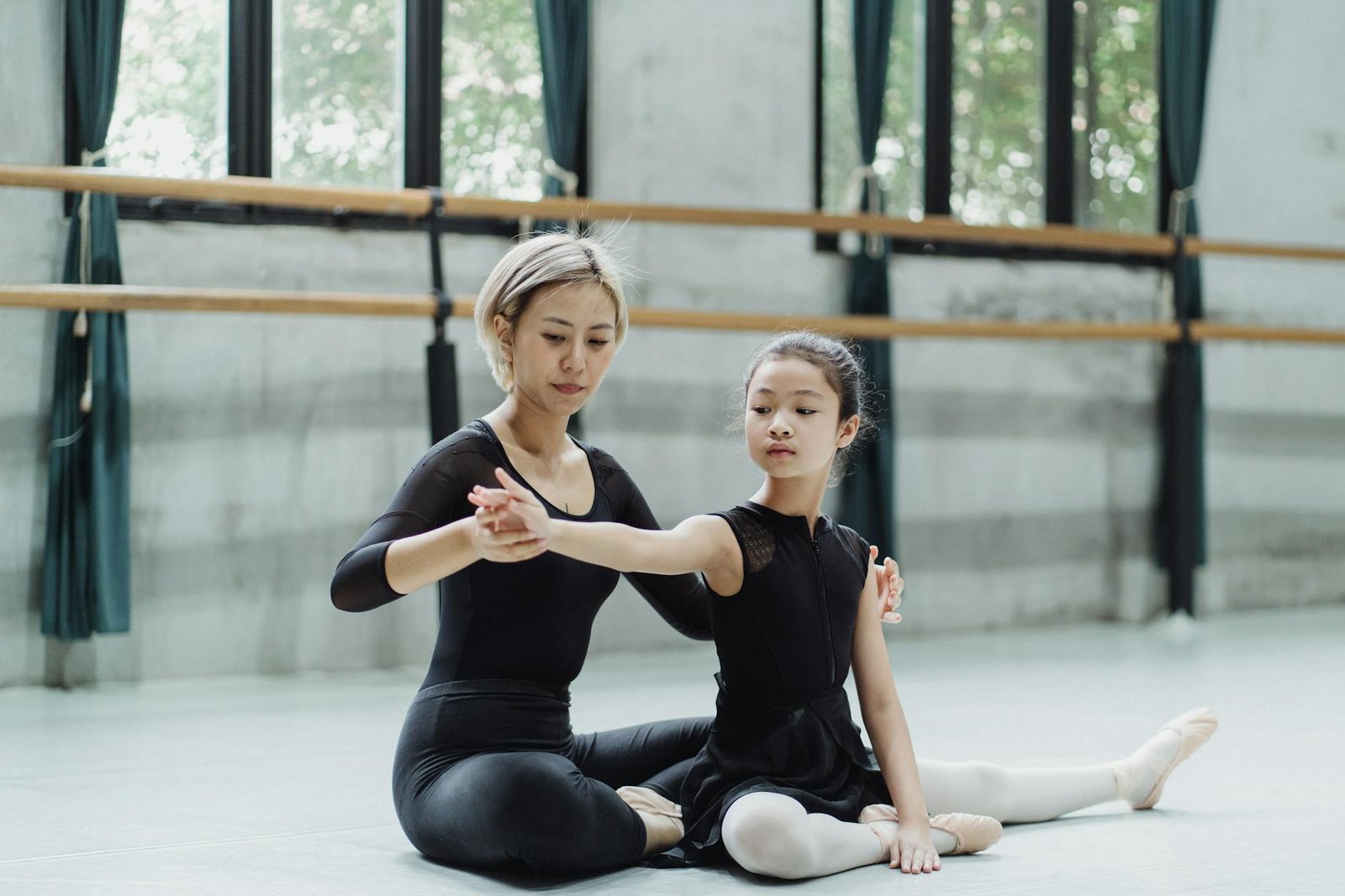 Young ethnic ballet teacher supporting arm of kid in uniform while exercising in dancing hall