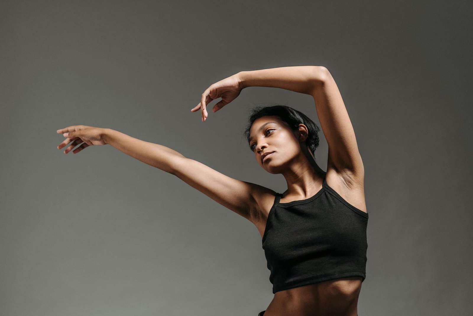 Woman in a dynamic dance pose with raised arms against a gray studio background.