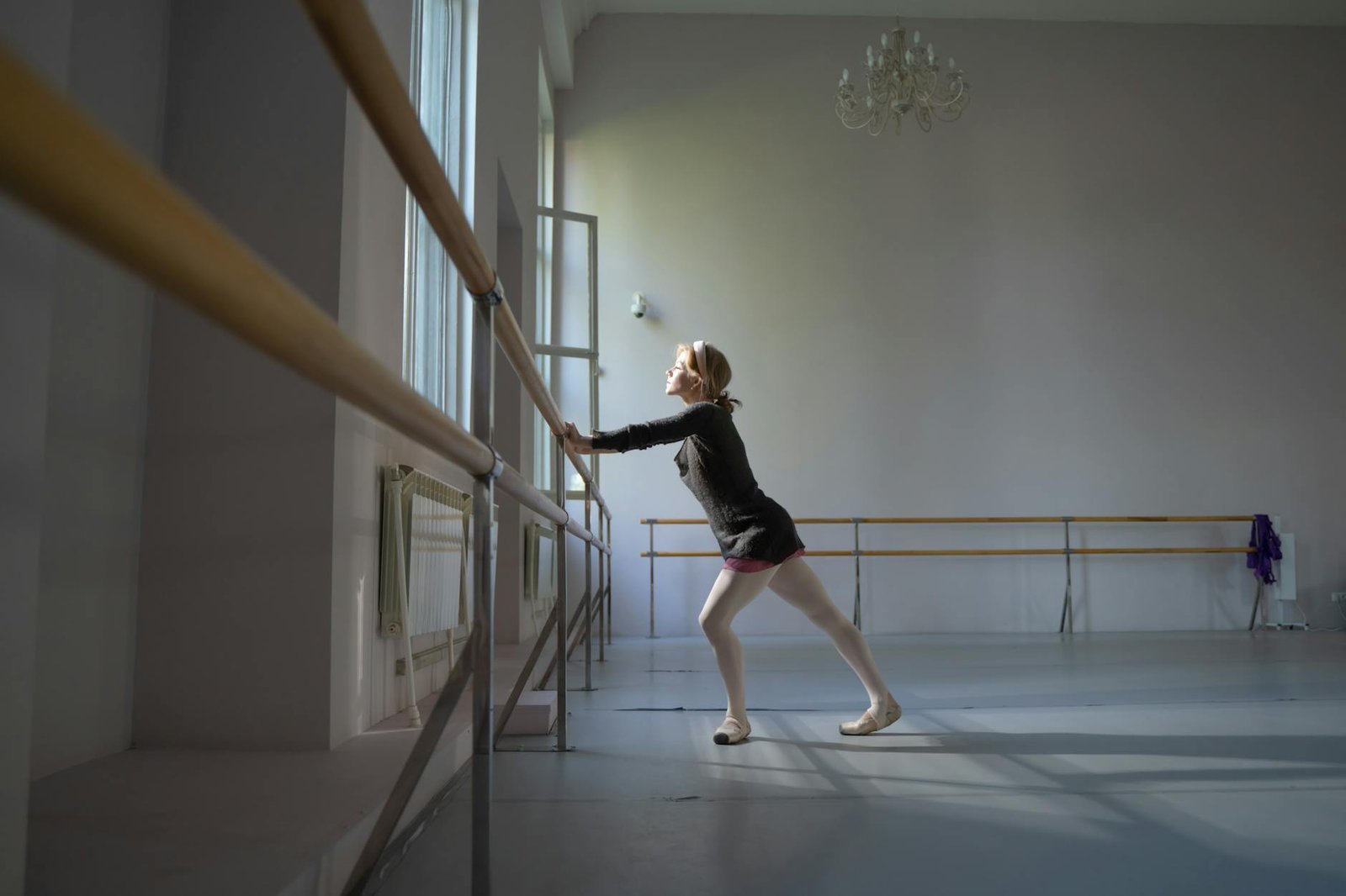 Young ballerina in dance studio practicing at barre, illuminated by soft natural light.