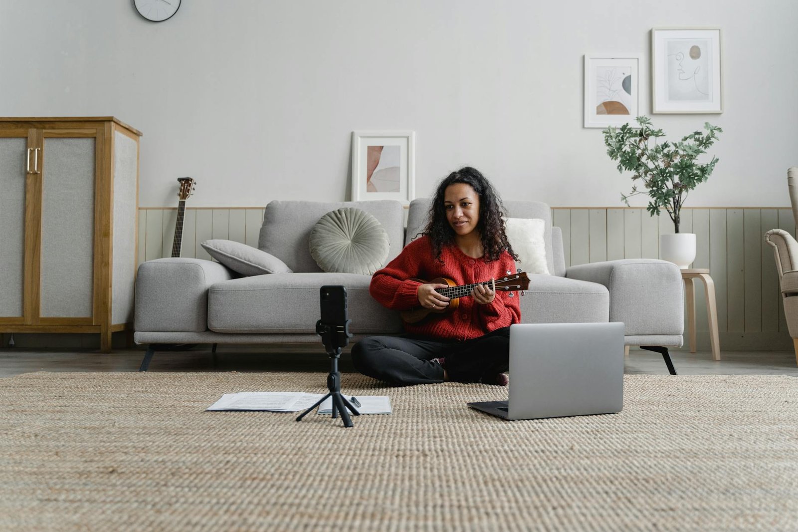 A woman sitting in her living room playing a ukulele during an online music lesson with a laptop and microphone.