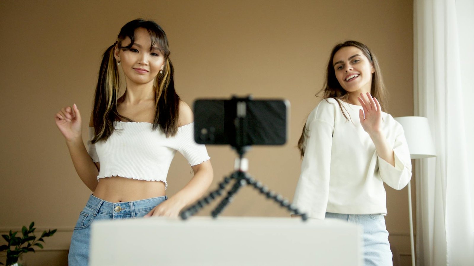 Two women joyfully dancing while recording with smartphone on tripod indoors.