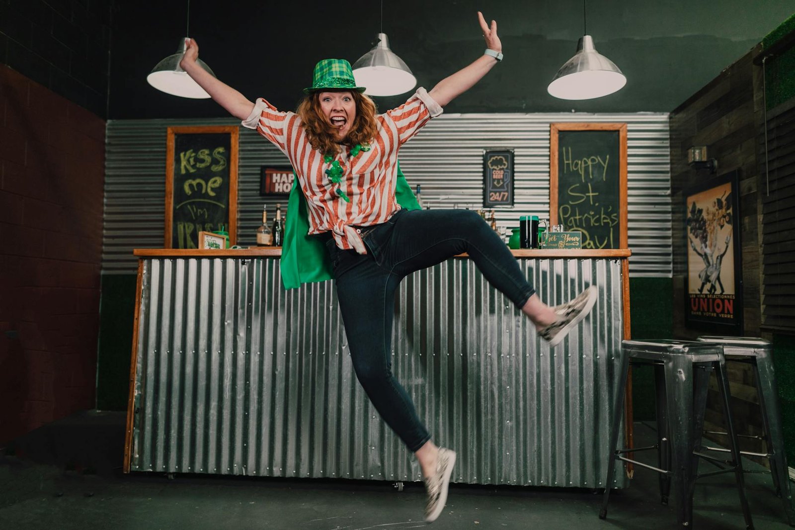 Woman in festive attire jumps joyfully in a bar decorated for St. Patrick's Day celebrations.