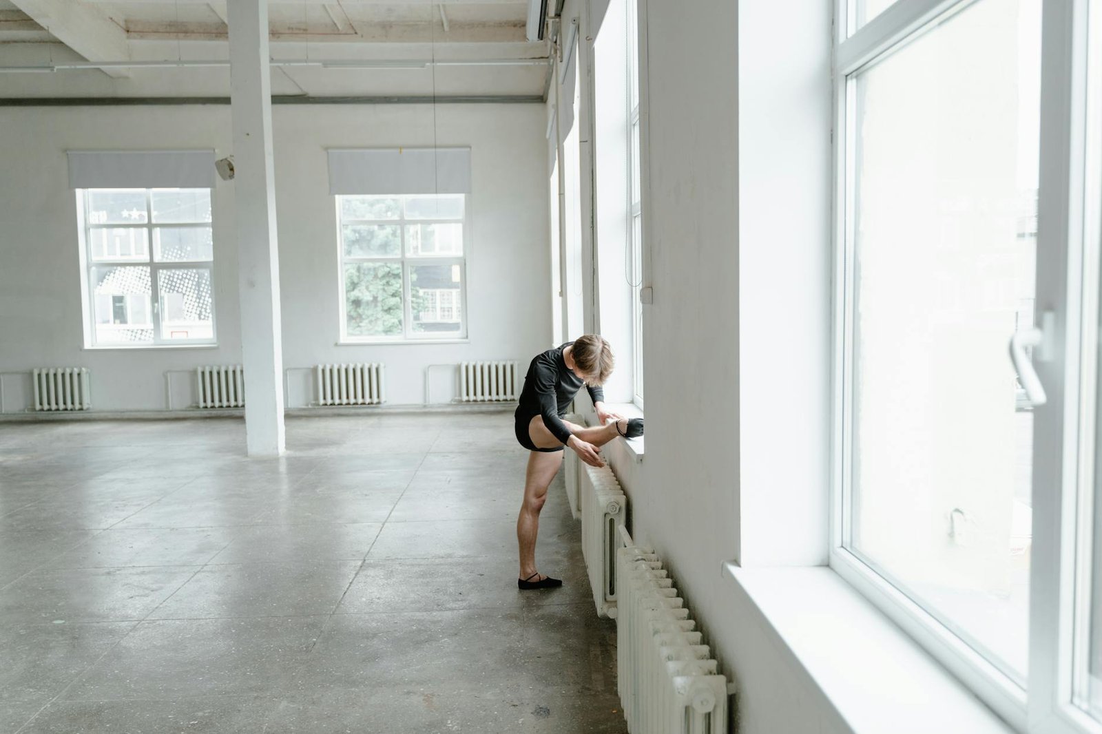 Ballet dancer stretching by a window in a large, bright studio space.