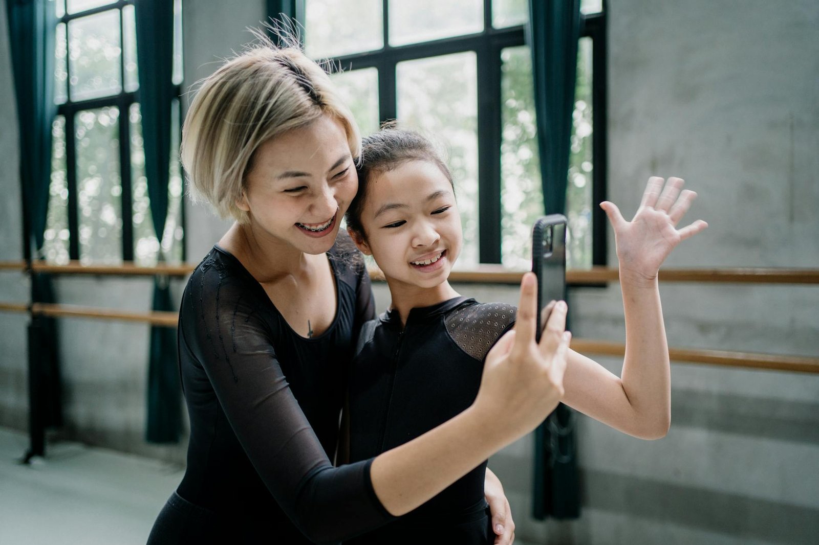 Delighted female ballet instructor and girl trainee wearing black leotards taking selfie on mobile phone in light ballet studio