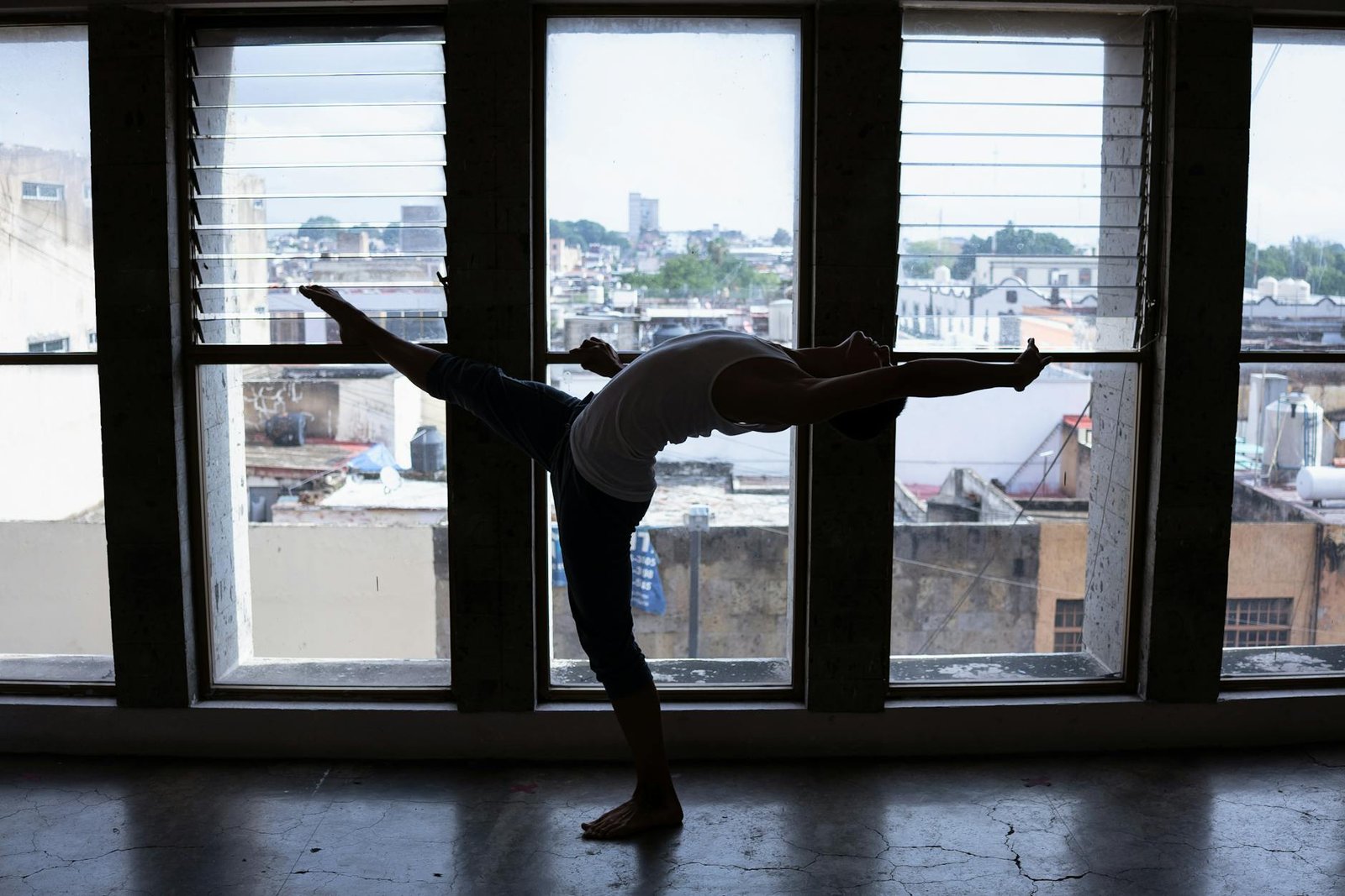 Backlit silhouette of a dancer performing a stretch in front of large urban windows, highlighting flexibility.