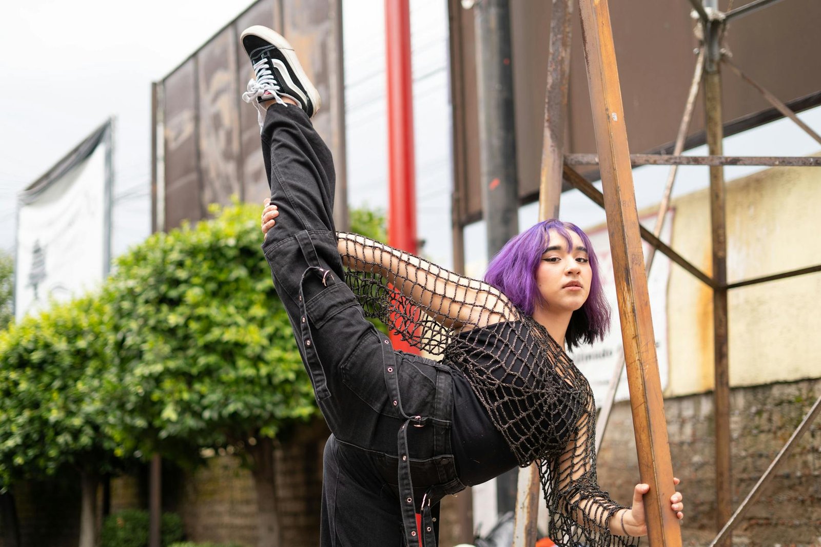 Confident street dancer with purple hair performs a high kick in an urban setting, showcasing flexibility and style.